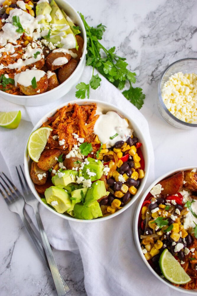 Three bowls with loaded burrito ingredients including shredded chicken, potatoes, avocado, corn, black beans, cheese, and crema, garnished with lime wedges and cilantro on a marble surface.