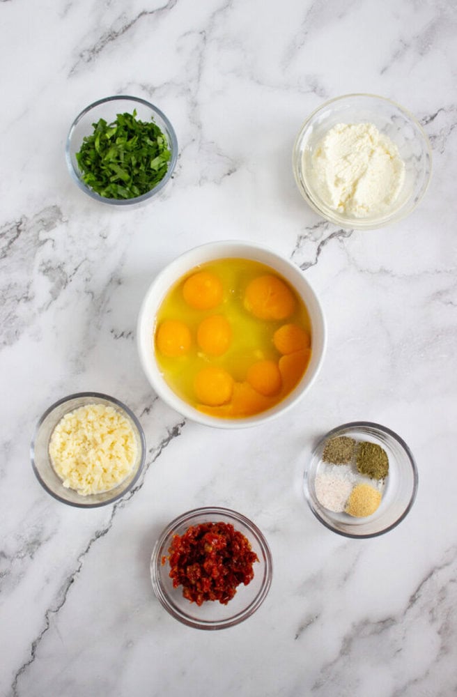 Six small bowls on a marble surface containing eggs, chopped herbs, ricotta cheese, shredded cheese, sun-dried tomatoes, and mixed spices.