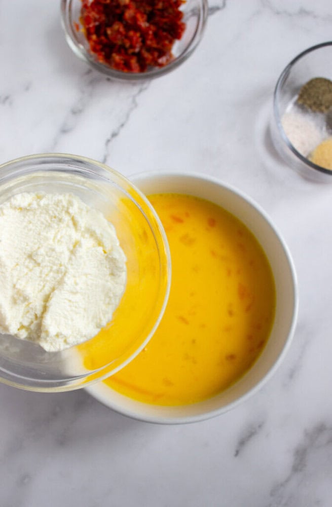 A bowl of beaten eggs next to a bowl of ricotta cheese, with small bowls of chopped sun-dried tomatoes and spices in the background on a marble surface.
