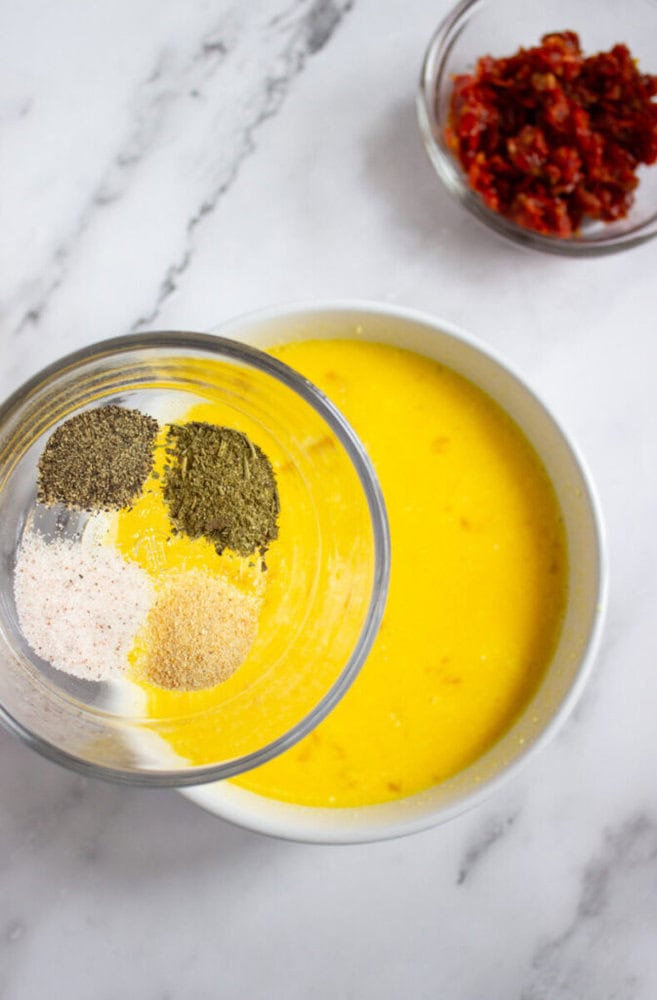 A glass bowl with black pepper, dried basil, salt, garlic powder, and onion powder held over a bowl of yellow liquid; chopped sun-dried tomatoes in a small bowl nearby.