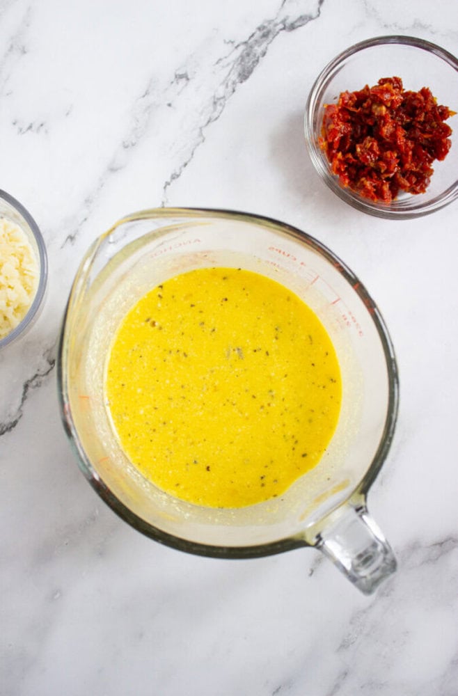 A glass measuring cup filled with a yellow liquid seasoning mixture, next to small bowls of shredded cheese and chopped sun-dried tomatoes on a marble surface.