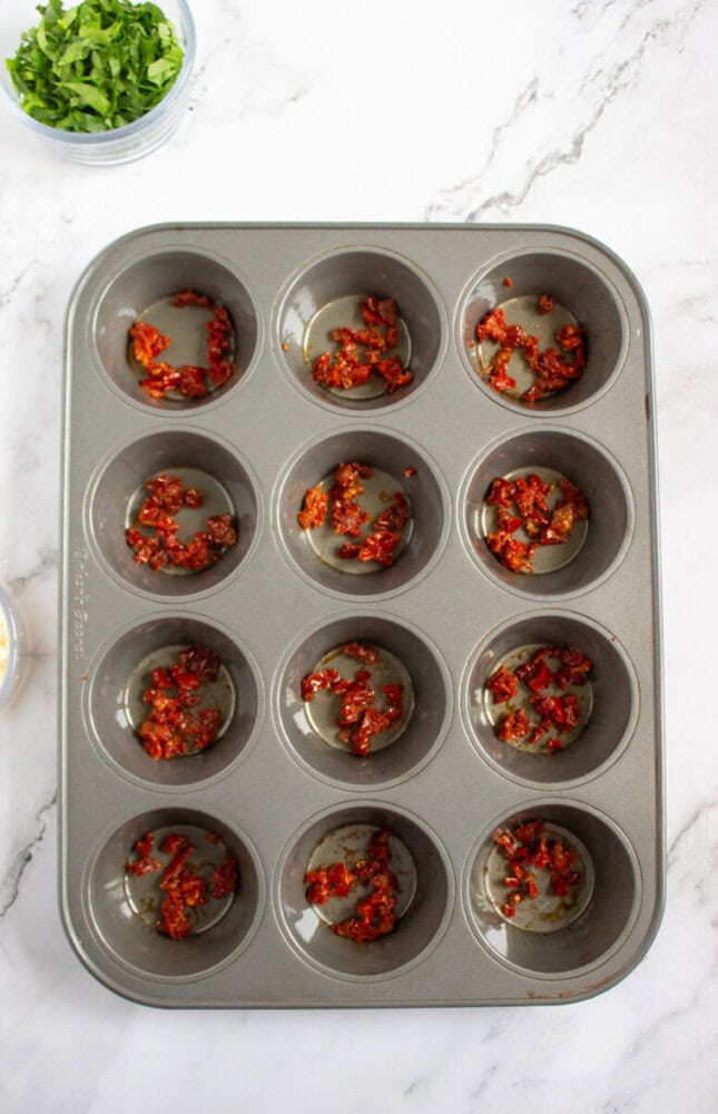 A muffin tin with several cups containing small amounts of chopped sun-dried tomatoes sits on a marble countertop; a bowl of chopped greens is in the corner.