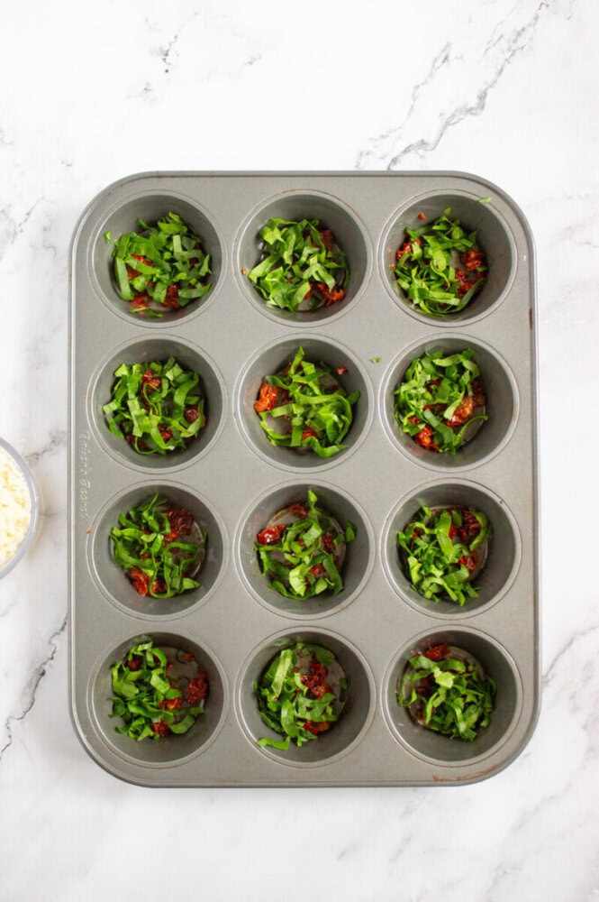 A muffin tin filled with chopped greens and sun-dried tomatoes in several cups, placed on a white marble surface.