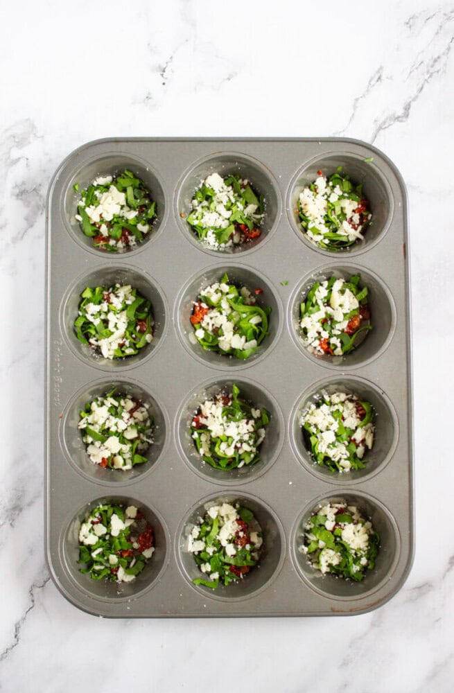 A muffin tin with twelve cups filled with a mixture of chopped vegetables and crumbled cheese, placed on a white marble surface.