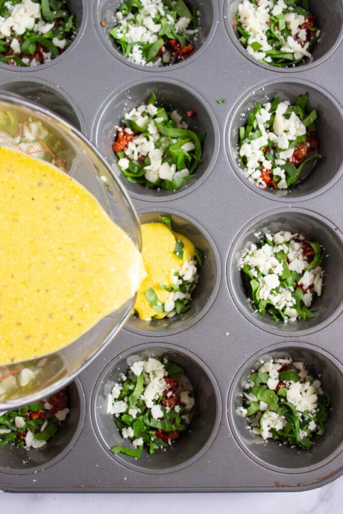 A muffin tin filled with chopped greens, sun-dried tomatoes, and cheese, with beaten eggs being poured into one of the cups.