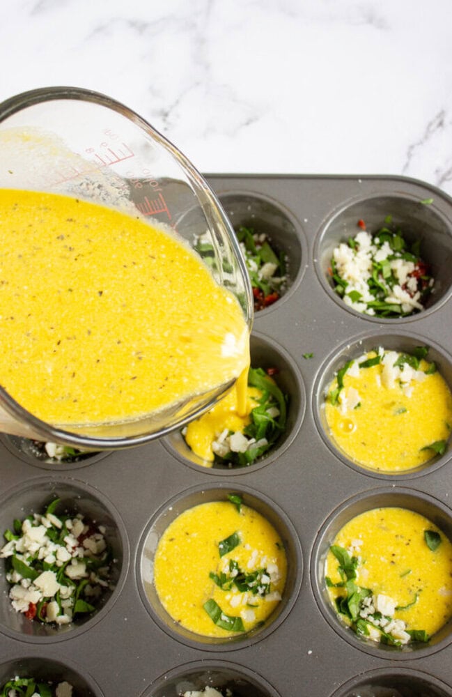 Egg mixture is being poured from a measuring cup into a muffin tin containing chopped vegetables and cheese, preparing for baking.