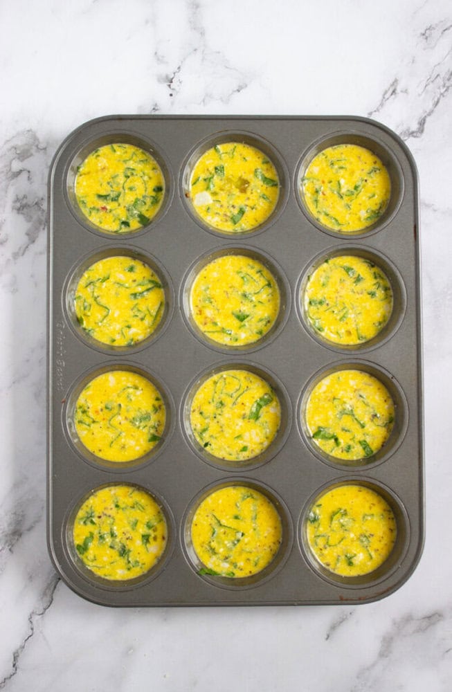 A 12-cup muffin tray filled with uncooked egg mixture containing chopped vegetables, placed on a marble countertop.