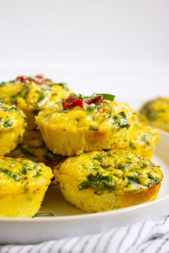 A plate of savory egg muffins with visible spinach, herbs, and pieces of tomato on top, arranged on a white surface.