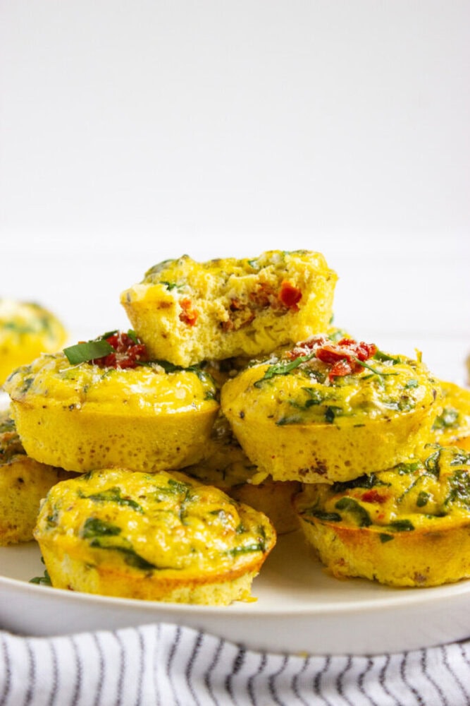 A plate of baked egg muffins with visible spinach and red pepper, stacked on top of each other, with one muffin showing a bite taken out.
