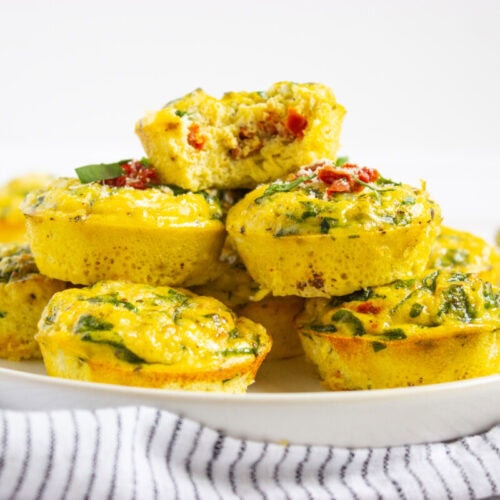A plate of baked egg muffins with spinach and diced vegetables, stacked in a pile with one missing a bite, on a white surface with a striped cloth nearby.