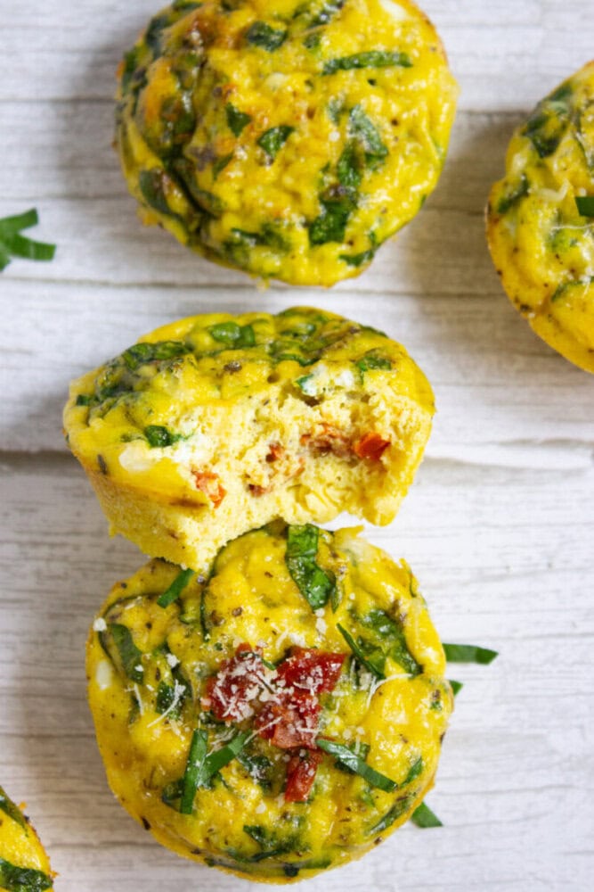 Three baked egg muffins with chopped spinach and sun-dried tomatoes, one with a bite taken out, arranged on a light wooden surface.