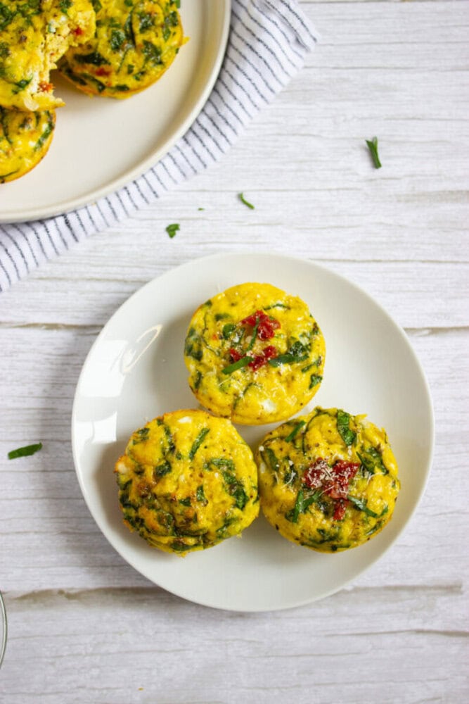 Three spinach and egg muffins are arranged on a white plate, with a striped napkin and more muffins visible in the background on a separate plate.