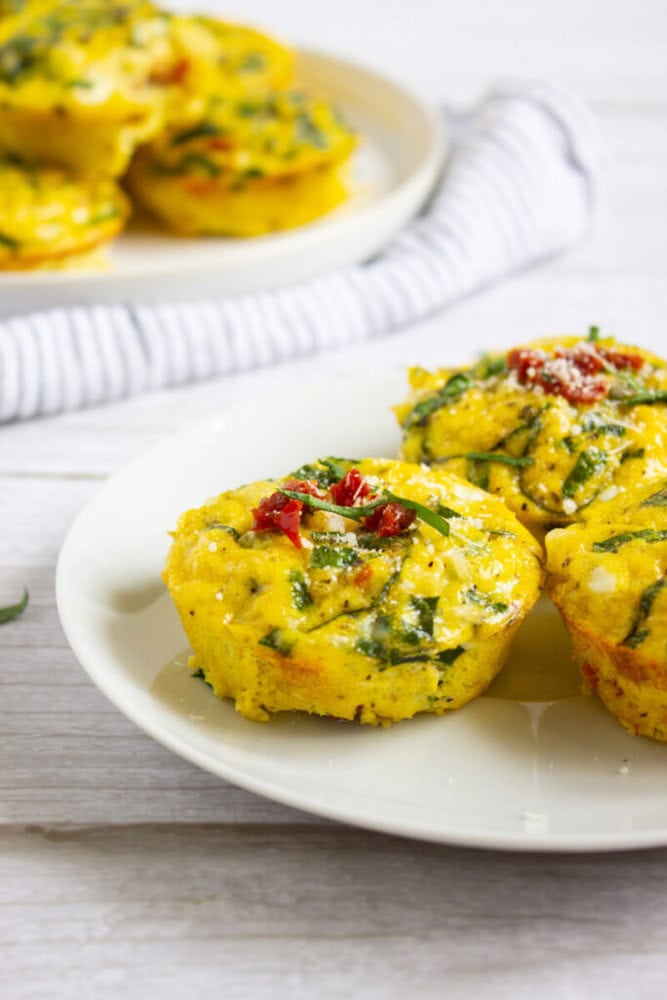 Two egg muffins with spinach and sun-dried tomatoes are served on a white plate, with more muffins in the background on another plate.
