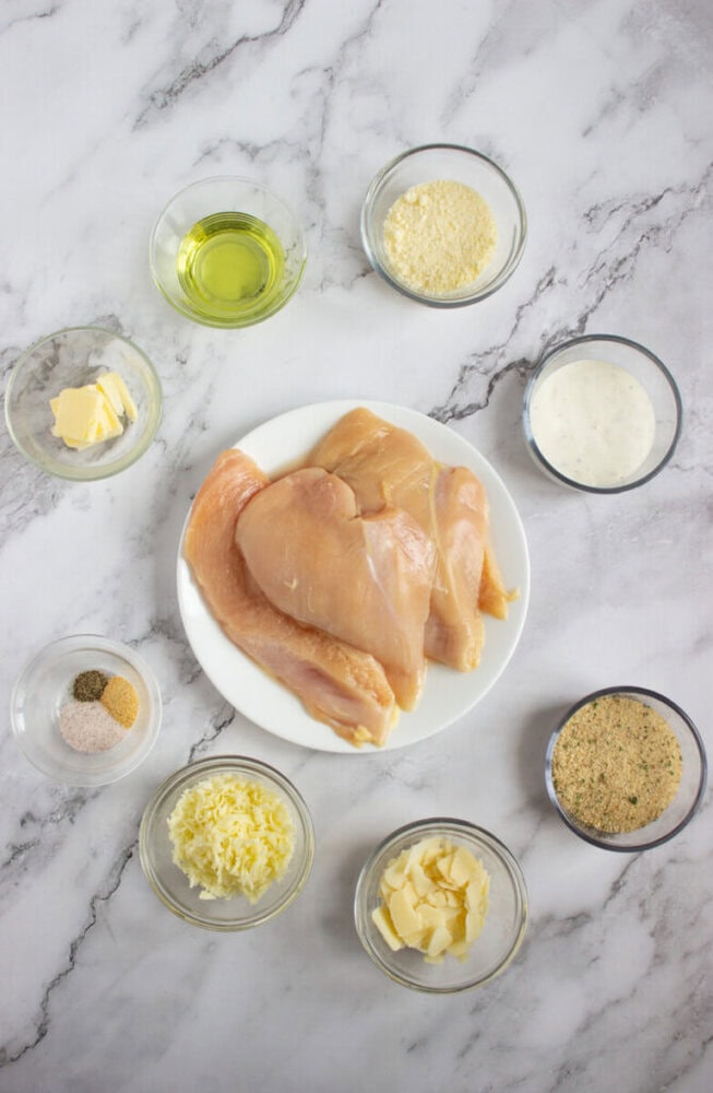 Raw chicken breasts on a white plate surrounded by small bowls of oil, cheese, butter, cream, seasonings, and breadcrumbs on a marble surface.