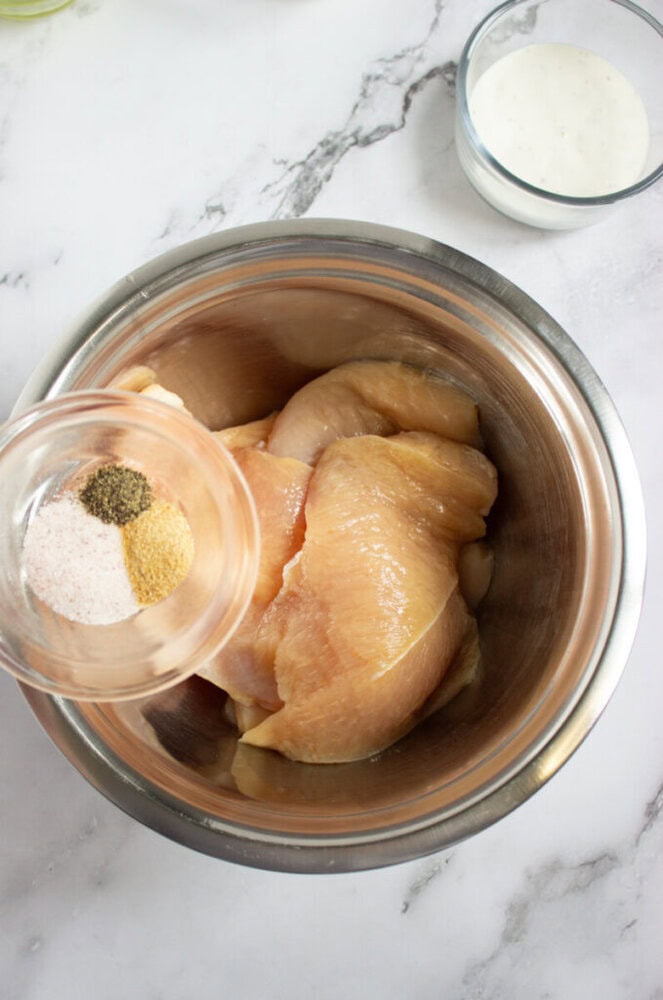 Raw chicken breasts in a metal bowl with a glass bowl of mixed seasonings being poured in; a small bowl of white sauce is in the background on a marble surface.