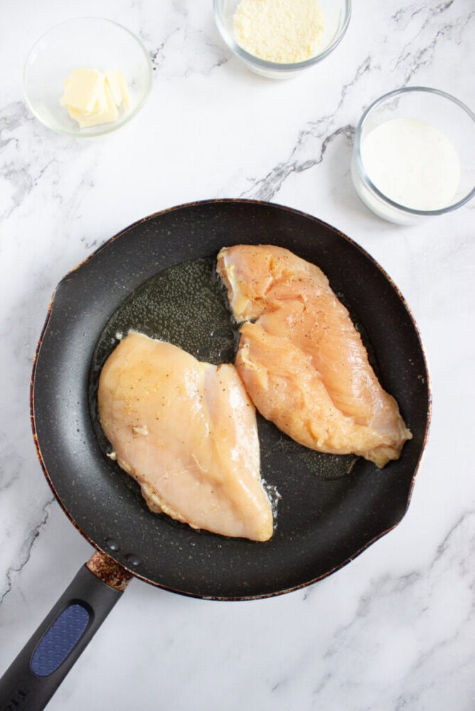 Two raw chicken breasts are cooking in a black frying pan. Nearby are small glass bowls containing butter, grated cheese, and a white sauce on a marble countertop.