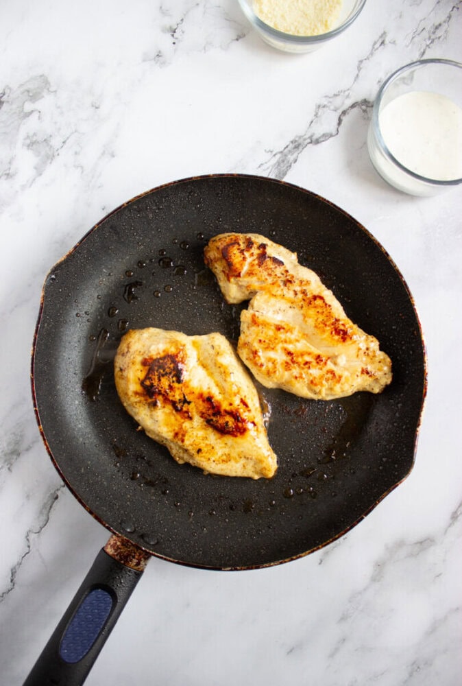 Two cooked chicken breasts in a nonstick frying pan on a marble countertop, with small bowls of grated cheese and white sauce nearby.