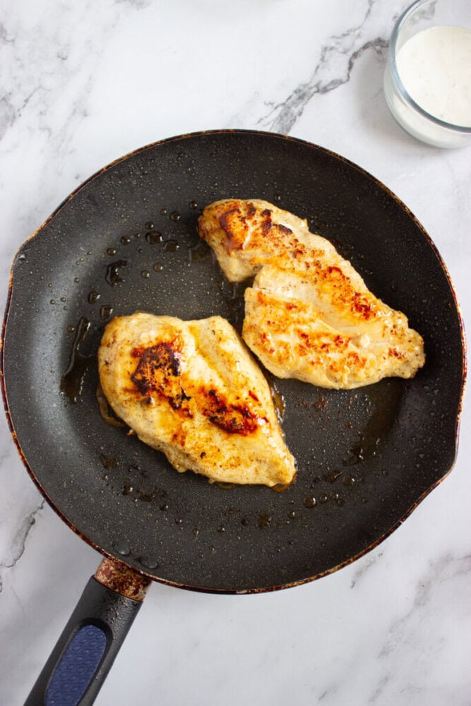 Two pieces of cooked chicken breast are in a black frying pan on a marble surface, with a small bowl of white sauce nearby.
