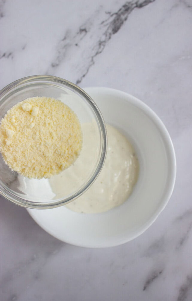 A glass bowl of grated cheese is held above a white bowl containing a creamy white sauce, on a marble countertop.