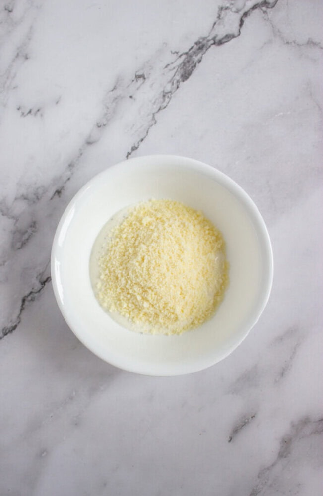 A white bowl containing a mound of powdered substance, likely grated Parmesan cheese, on a marble surface.