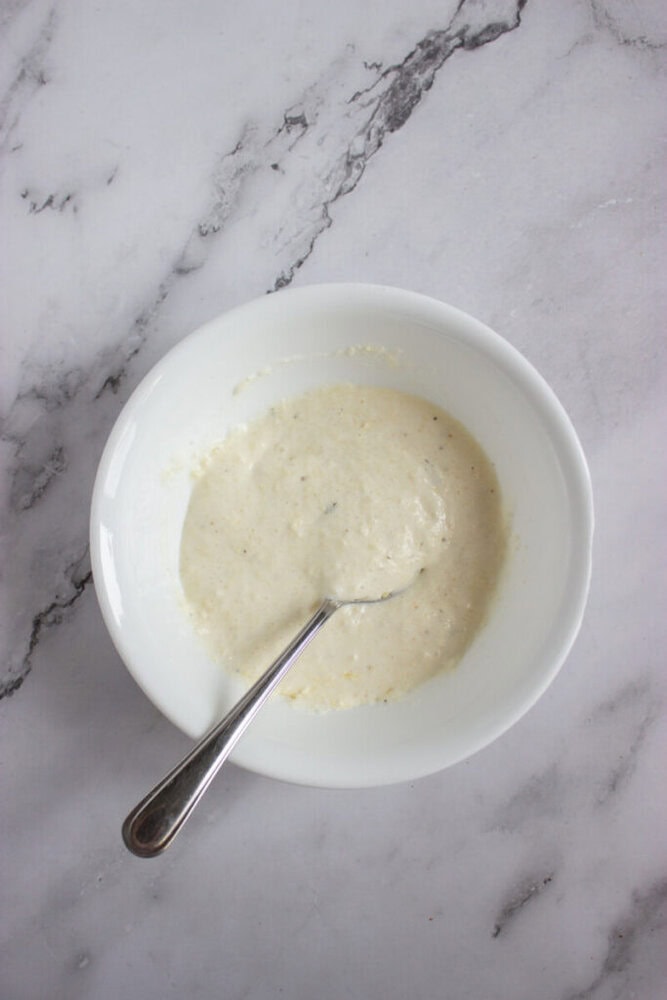 A white bowl with a spoon inside contains a creamy, pale mixture on a gray marble surface.