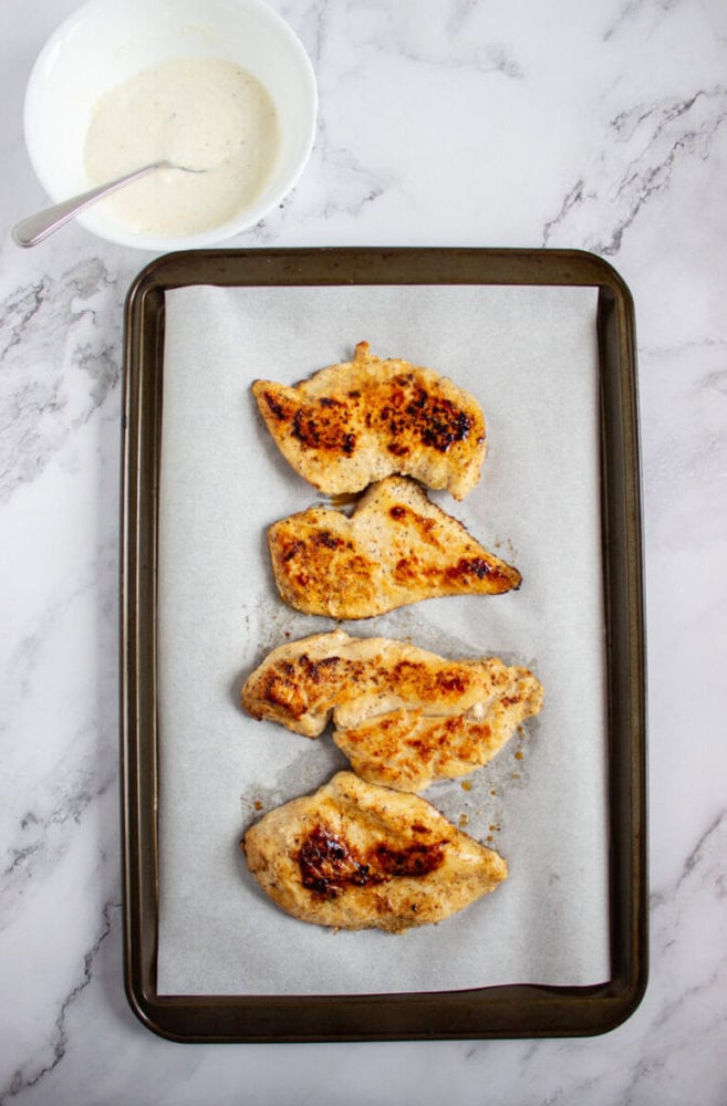 Four cooked chicken breasts on a parchment-lined baking tray, with a bowl of creamy sauce and a spoon in the top left corner on a marble surface.