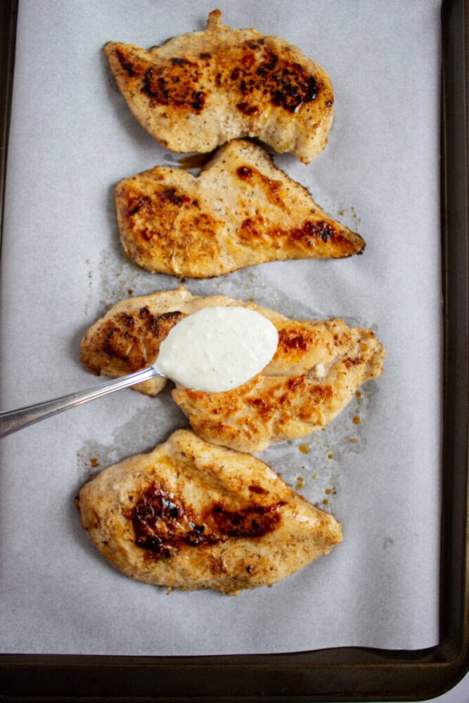 Four cooked chicken breasts on parchment paper, with a spoon spreading a creamy white sauce on one piece.
