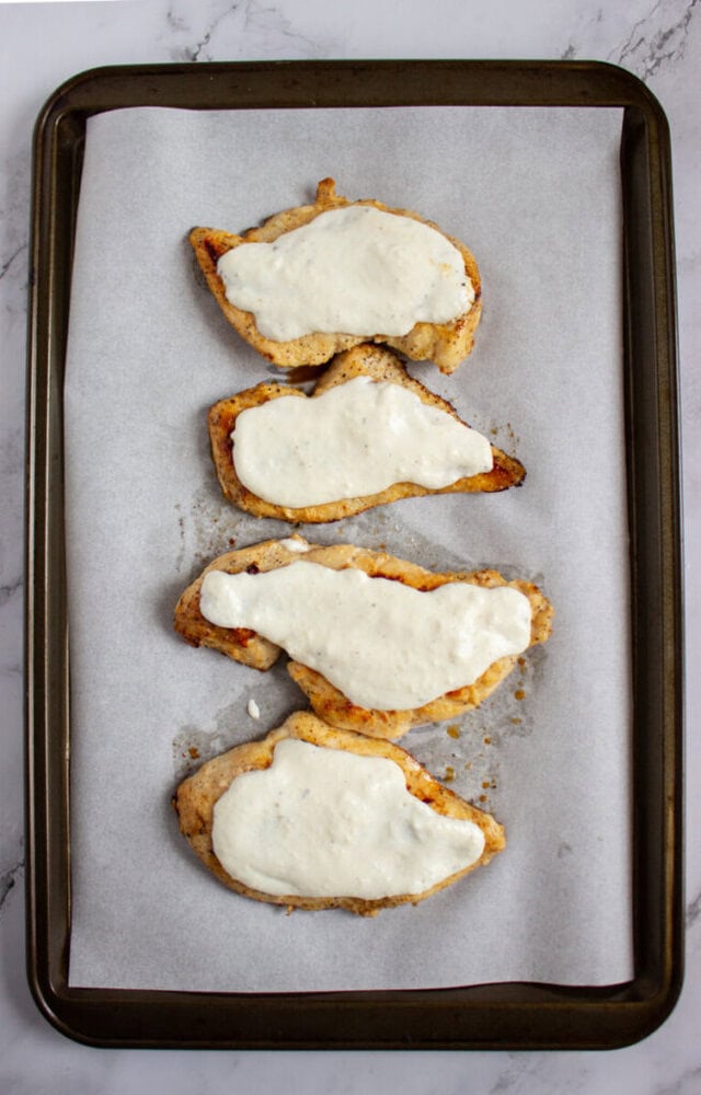 Four baked chicken breasts topped with melted white cheese, arranged in a row on parchment paper on a baking sheet.