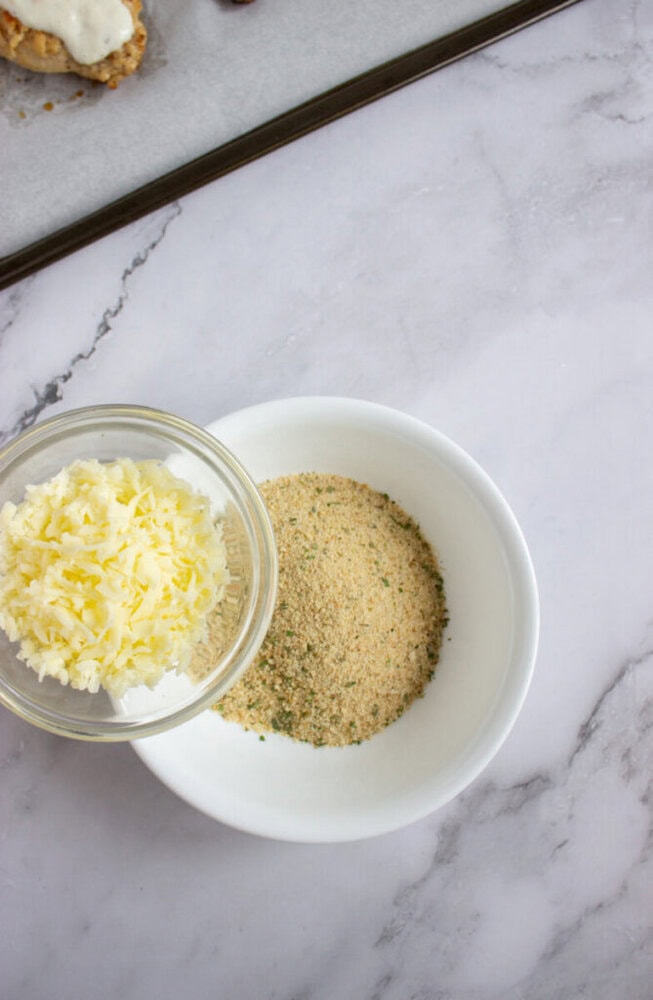 A bowl of grated cheese next to a bowl of seasoned breadcrumbs on a marble countertop.