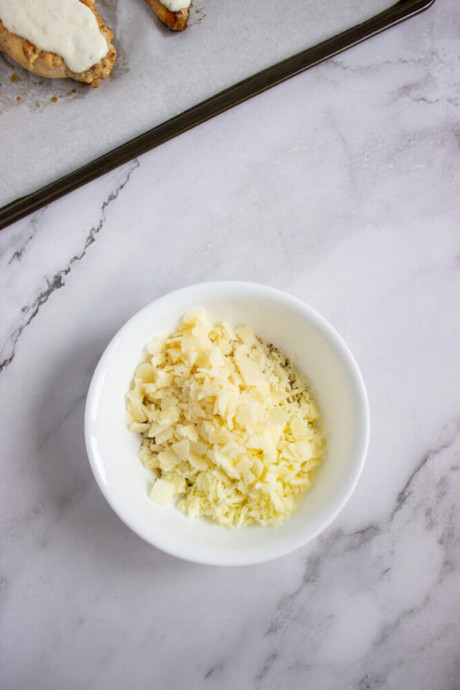 A white bowl filled with grated and cubed cheese is on a marble surface, next to a baking tray with partially visible food.