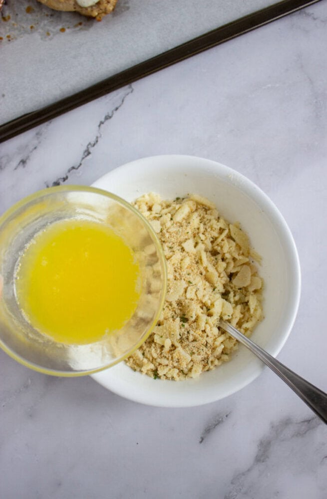 A bowl of bread crumb mixture with a spoon sits beside a smaller bowl of melted butter on a marble surface.