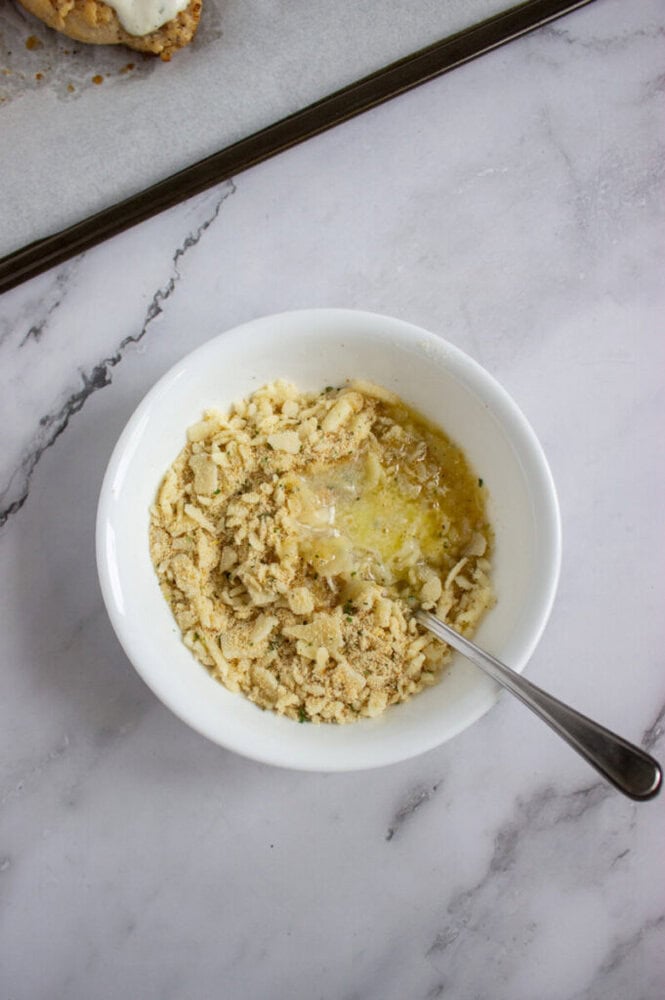 A white bowl containing crushed crackers, melted butter, and seasonings with a metal spoon on a marble countertop.