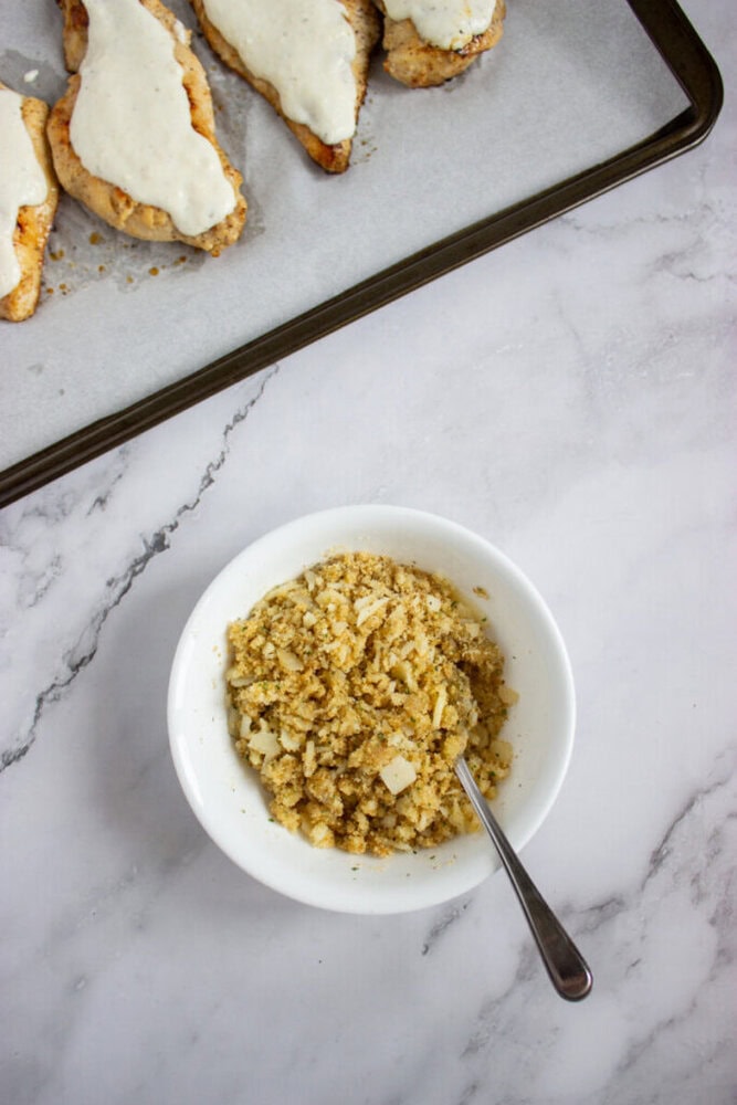 A white bowl of breadcrumb mixture with a spoon sits on a marble counter next to a tray of partially prepared chicken breasts topped with melted cheese.