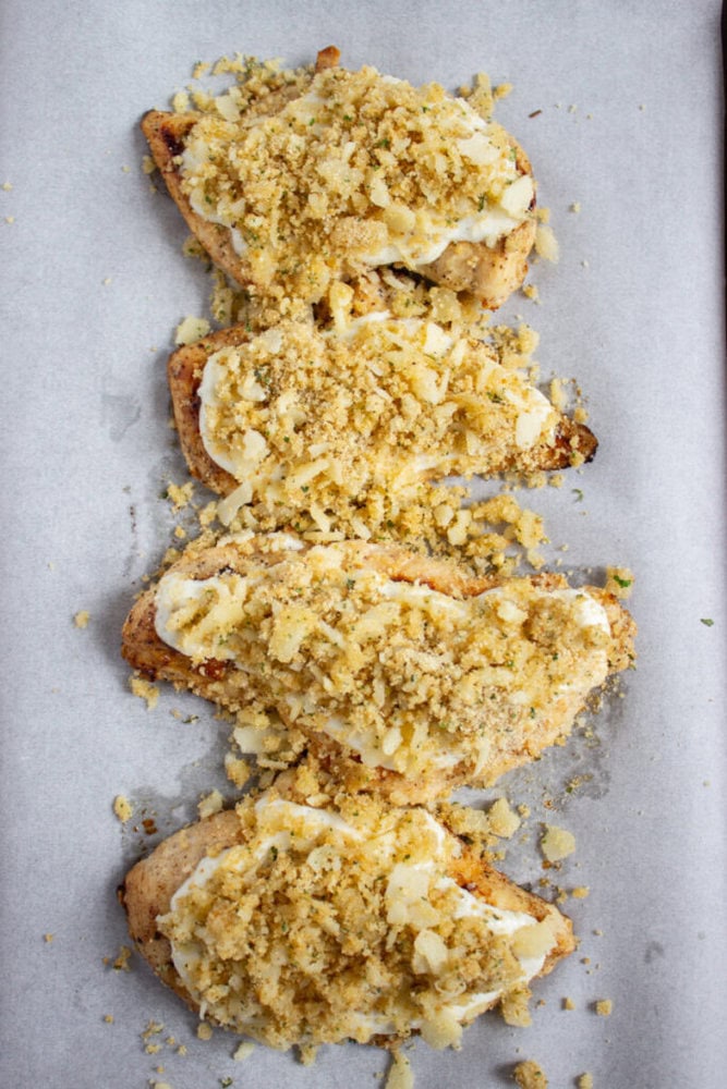 Four baked chicken breasts topped with a creamy sauce and a layer of crushed breadcrumbs, arranged in a row on a sheet of parchment paper.