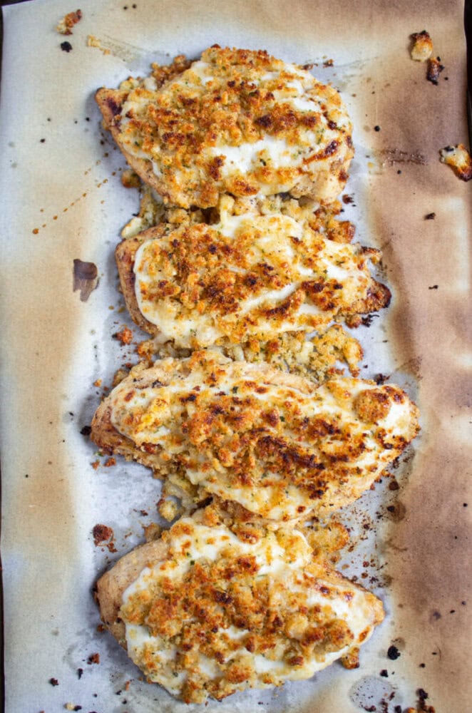Four baked chicken breasts topped with a golden, crispy cheese and breadcrumb crust on a sheet of parchment paper.
