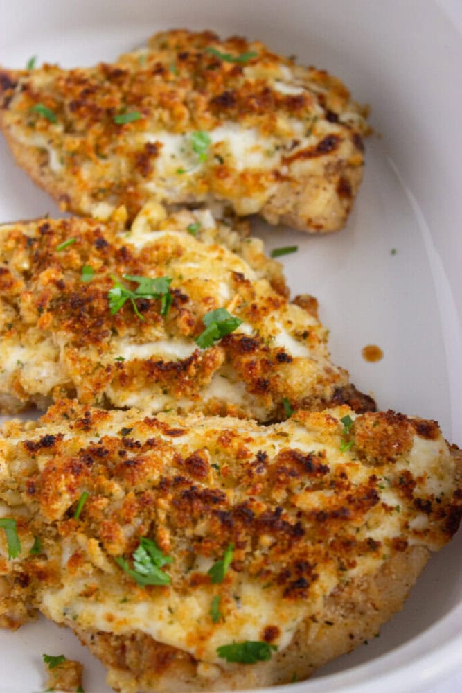 Three pieces of baked chicken breast topped with a golden breadcrumb and herb crust, garnished with chopped parsley, in a white dish.