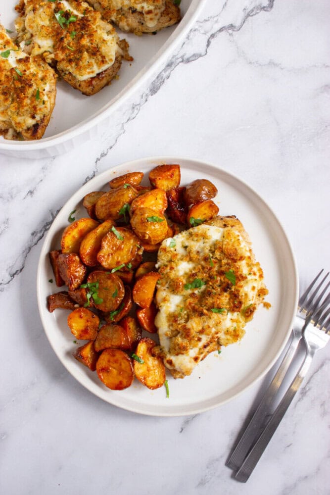 A plate of breaded baked chicken breast with melted cheese and a serving of roasted potatoes, garnished with herbs, next to a fork and knife on a marble surface.