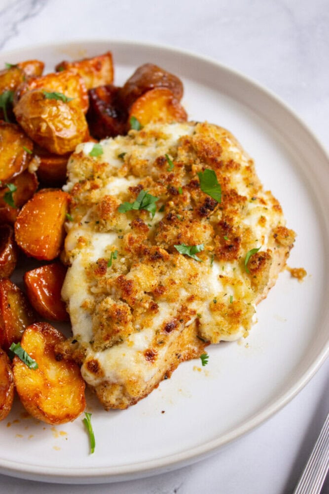 A breaded chicken breast with melted cheese and herbs is served next to roasted potatoes on a white plate.