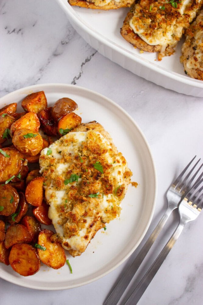 A plate with baked chicken breast topped with melted cheese and breadcrumbs, served alongside roasted potatoes. Two forks are placed next to the plate on a marble surface.