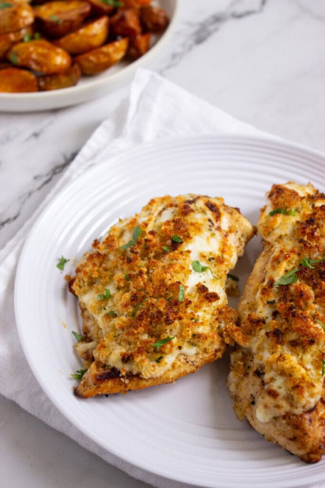 Two pieces of baked chicken breast topped with a golden, cheesy breadcrumb crust on a white plate, with a bowl of roasted potatoes in the background.