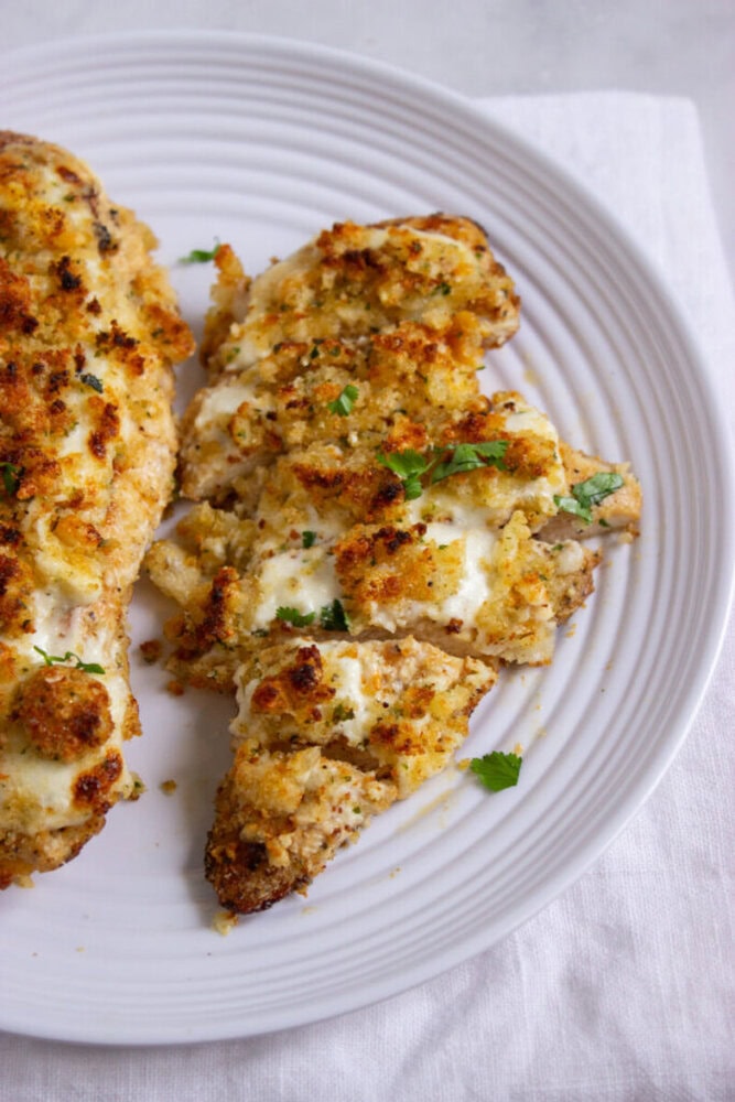 Breaded, baked chicken breast topped with melted cheese and herbs, sliced and served on a white plate.