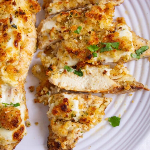 Sliced baked chicken breast topped with melted cheese and breadcrumbs, garnished with chopped parsley, served on a white plate.