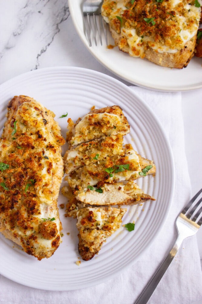 A plate with breaded, baked chicken breast sliced and garnished with herbs. Another plate with more chicken pieces and a fork is visible in the background.