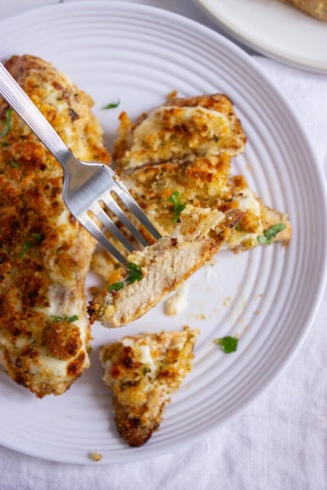 A fork holds a piece of breaded, baked chicken breast over a white plate with more pieces of chicken and some parsley garnish.