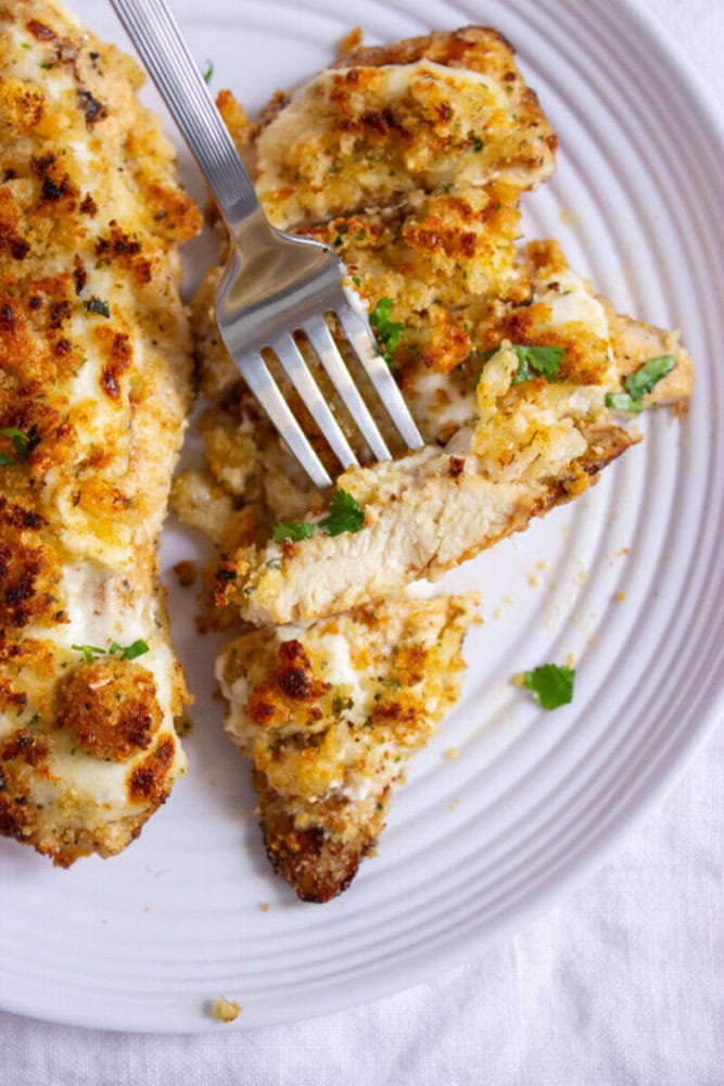 A fork cutting into a piece of breaded and baked chicken breast on a white plate, garnished with parsley.