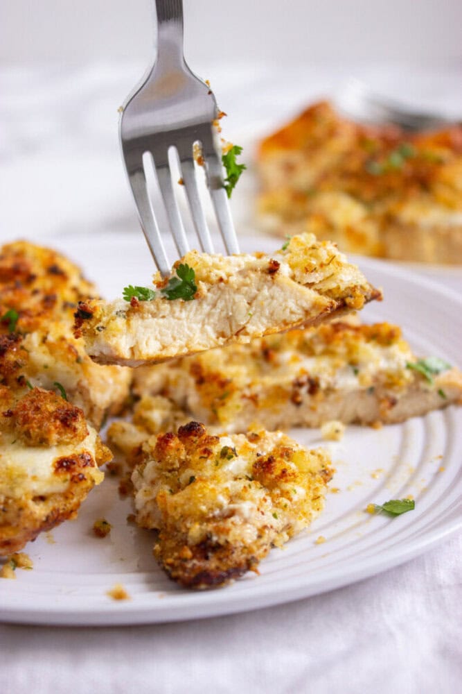 A fork holds a sliced piece of baked, breaded chicken garnished with herbs on a white plate with more chicken pieces in the background.