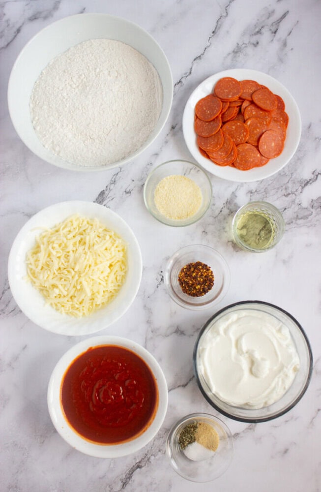 Bowls of pizza ingredients on a marble surface, including flour, pepperoni, shredded cheese, tomato sauce, Parmesan, spices, and a large bowl of white yogurt or cream.