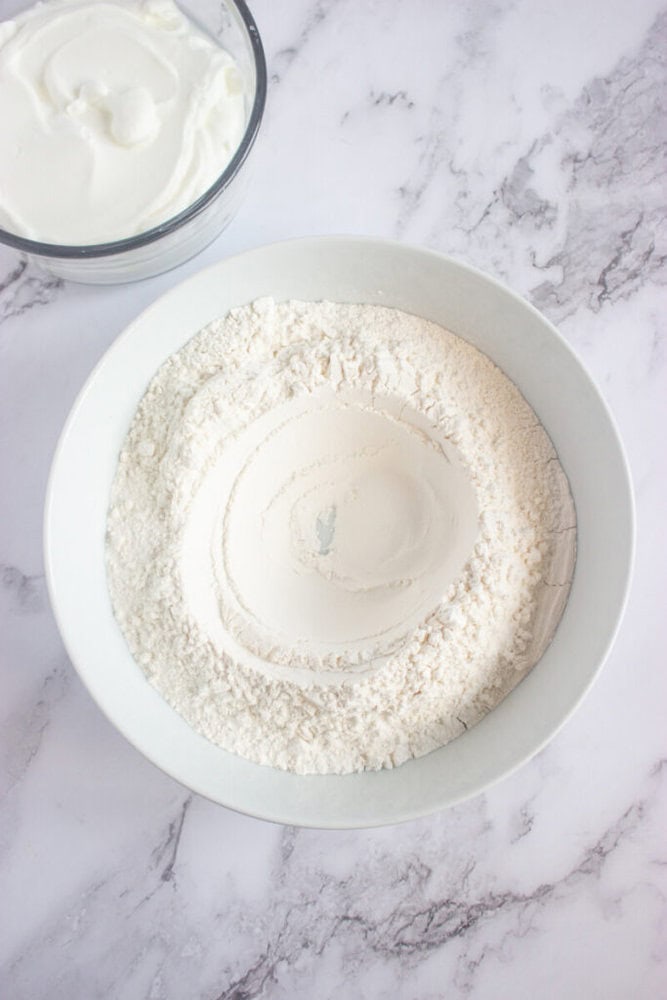 A white bowl filled with flour sits on a marble surface next to a smaller bowl containing thick white yogurt.