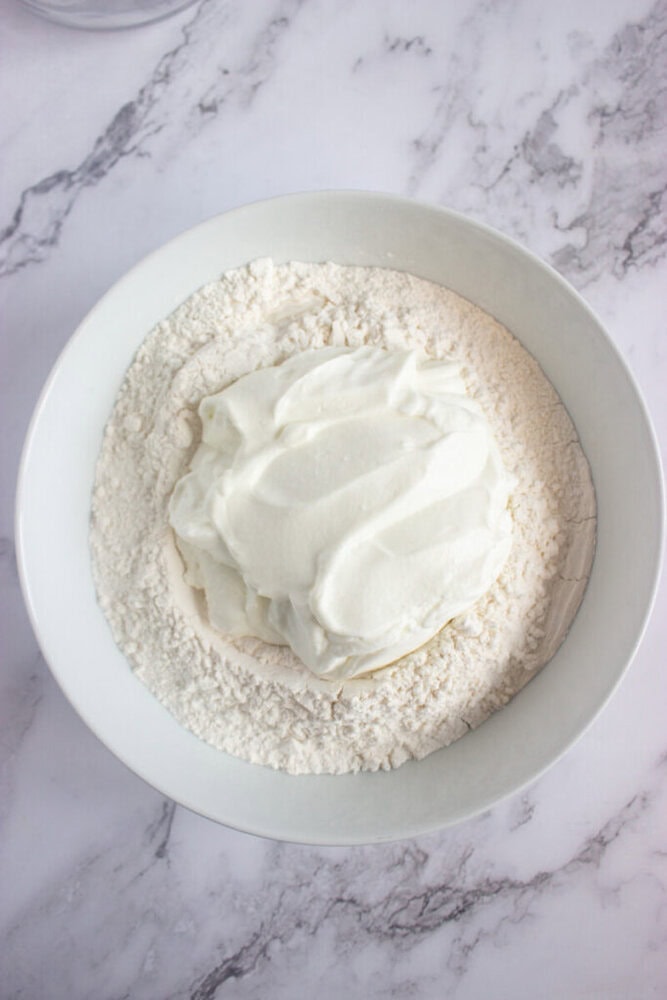 A bowl containing flour with a large dollop of plain yogurt on top, placed on a marble countertop.