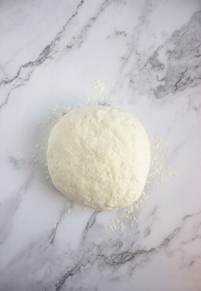 A ball of raw dough sits on a lightly floured, white marble surface.